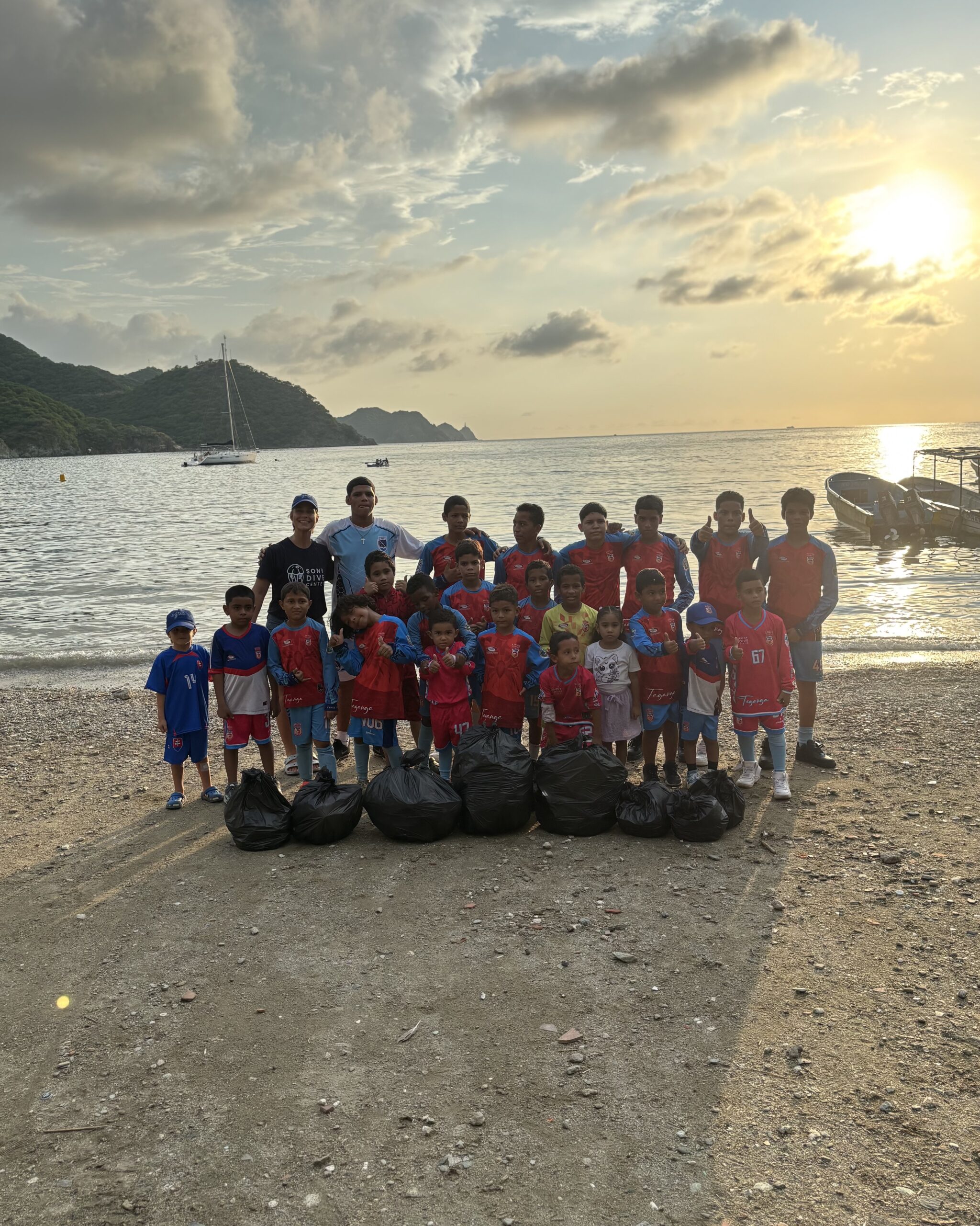 Group of local children and volunteers posing with collected trash bags after a beach cleanup in Taganga, Santa Marta at sunset.